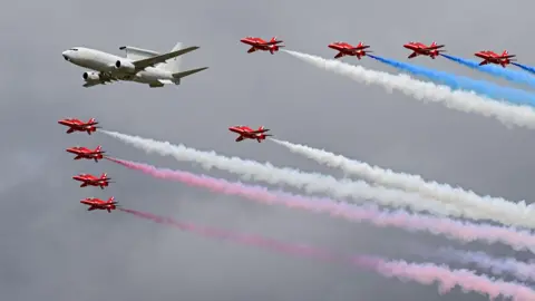 Getty Images The Red Arrows flying in formation, with red and white smoke trailing behind. Also flying alongside them is a larger white jet-style plane.