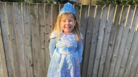 Grace is wearing a light blue dress and is smiling as she stands in front of a wooden fence. 