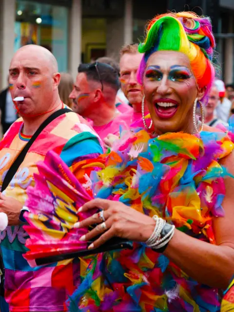 A Pride parade participant wearing a brightly coloured rainbow wig, large hoop earrings and a multicoloured feathered outfit holds a pink folding fan while walking through a crowded city street parade.
