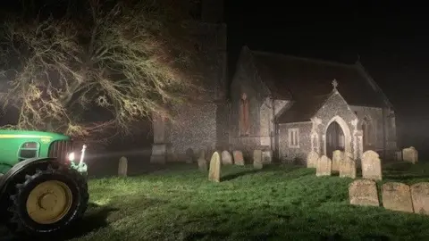 Matthew Welch A green tractor shines its light through the darkness on the entrance of a church. The gravestones in the churchyard with green grass are also visible. There is tree behind the tractor.