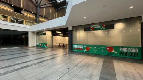 Tom Jackson/BBC General view of the exterior of the ping pong parlour. It is on the ground floor of a shopping centre with a "Ping Pong parlour" sign outside and a green banner with "Step inside & Play for Free" on it.