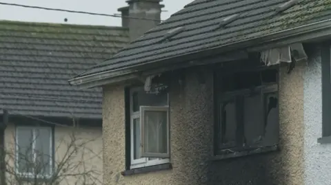 The camera is zoomed in on the top two windows of a light brown house. The window on the right id smashed and is black with burn marks. The area around the window is also stained black. The window to the right is open and relatively untouched by heat. 