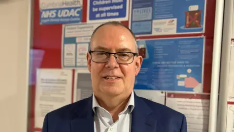Dave is wearing a smart navy suit and is pictured in front of the noticeboard at the urgent dental access centre in Carlisle. He is wearing glasses and has very short dark hair.