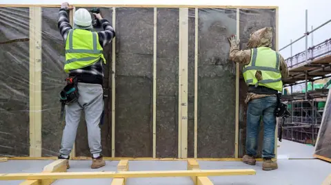 Two workers in high-viz jackets stand with their backs to the camera holding up a partition wall, the worker on the left is holding a drill in their right hand and is drilling into the wall.