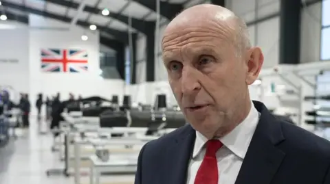 Defence Secretary John Healey stands in a factory. He has a dark suit with a white shirt and red tie on.