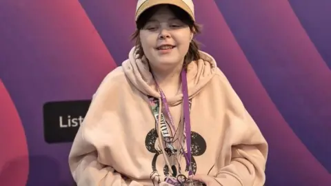 Lilly in a radio studio. She is wearing lanyards. She has a beige Micky Mouse hoody on and a beige hat.