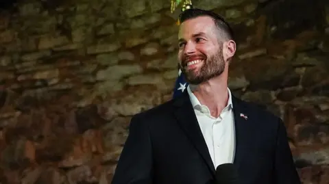 Clay Fuller, in a suit jacket and white collared shirt, looks to the side and smiles