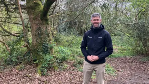 James Wright from the Dartmoor National ParkAuthority, standing in the Wray Valley campsite. He is in a clearing, with brown fallen leaves on the grouns and moss covered trees behind. He is smiling and wearing beige trousers and a dark blue zip fleece. He has a beard and short hair that is greying.