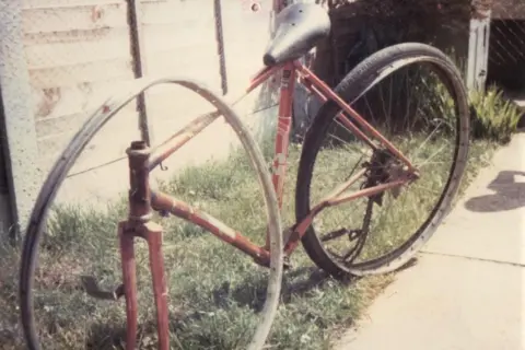 Mark Williams An old photo from 1982 of Mark Williams' damaged bicycle. It is a red road bike with a slightly damaged back wheel and the chain broken. The front wheel is missing, and the metal wheel rim with no spokes is leaning up against the frame of the bike. 