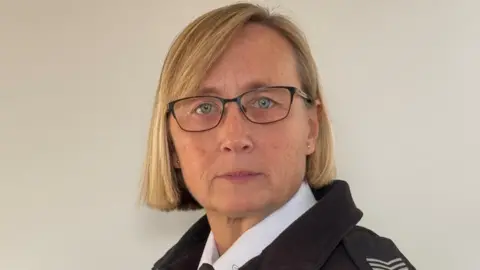 Laura Heggie, who has short blonde hair and wears black rimmed glasses. She is wearing her police uniform, consisting of a white shirt and a black coat that has epaulettes on.