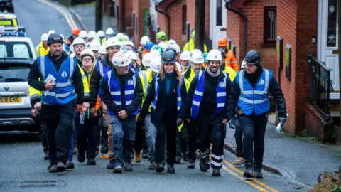 South Shore A large group of  workers in hard hats and high-vis clothing walk up a residential street together. Several people at the front wear blue safety vests; a woman with long blonde hair can be seen in the centre of the front row. Terraced brick houses line the right side of the street, and parked vehicles are visible on the left.