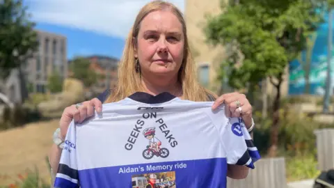 BBC/Simon Thake A woman with blonde hair holds up a white and purple cycling shirt with a picture on the front of Aiden Batchelor.