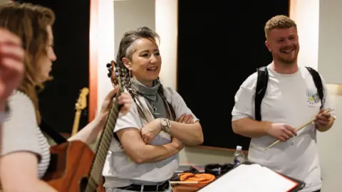 Alistair More KT Tunstall in white T-shirt and scarves is standing in a bare rehearsal room with Jamie Rea, also in white T-shirt. They are surrounded by musicians.