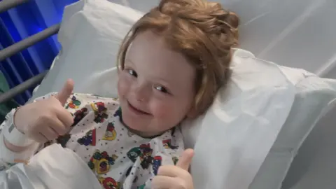 Family handout Phoebe is laying on a hospital bed with white sheets. She has ginger hair in a ponytail and has a bandage on her arm. She is wearing patterned pyjamas and has her thumbs up.