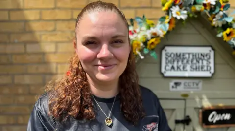 Charley Coleman-Pollard with her cake shed in the background which is decorated with flowers and has and open sign on the door. Charley is wearing a dark coloured top with a necklace.
