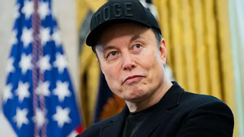 AFP via Getty Images Elon Musk wearing a black jacket a baseball cap. The US flag hangs in the background.
