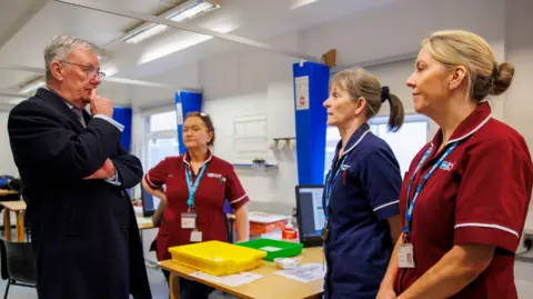 Liam McBurney/PA Wire Health Minister Mike Nesbitt talks with three female healthcare staff at the Ulster Hospital Vaccination Centre in Belfast. He is resting one hand on his chin as he listens to the women who are all dressed in health service uniforms.
