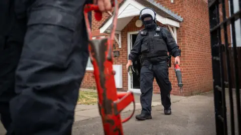 Merseyside Police Two police officers (one partly in shot) in riot gear with a battering ram after leaving a house after executing a warrant