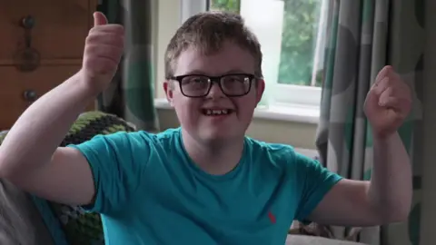 A teenage boy with Down's syndrome wearing a blue T shirt holds both thumbs in the air