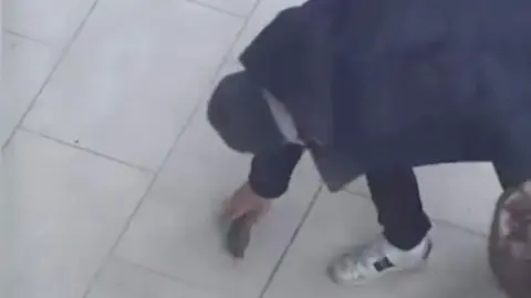 PA Media CCTV still of a man crouching on a tiled pavement as he places a small cylindrical object on the ground. He is wearing a dark coat and white trainers.