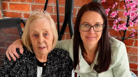 Two people are seated closely together indoors against a red brick wall. The person on the left has short light-colored hair and wears a dark patterned jacket and a decorative headband, while the person on the right has long dark hair, wears glasses, and is dressed in a light-colored top. The person on the right has one arm around the other’s shoulder. In the background, there is black metal furniture and a branch with pink artificial flowers.