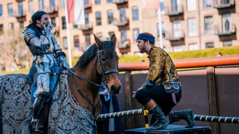 Josh Heely, Royal Armouries A jouster is sitting on a horse wearing full armour. She is shocked as a man with her hand covering her mouth as a man also wearing 16th century clothing is on one knee and proposing to her. 