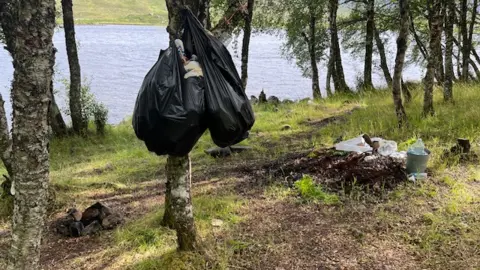 Highland Council A number of full bin bags are hanging from a tree in a wooded area next to a loch. There is more litter on the ground. 