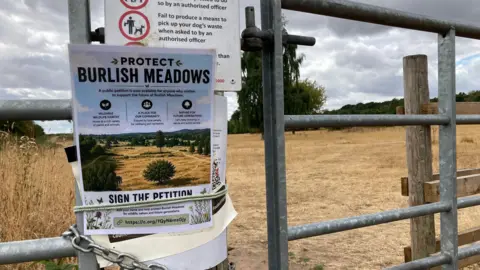 A poster that reads "protect Burlish Meadows" is pinned to a metal gate in front of a large open field that has turned brown from the dry weather.