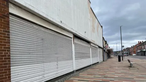 Simon Thake/BBC A deserted high street. The nearest shop is closed permanently with four shutters pulled down and white paint peeling from the signage