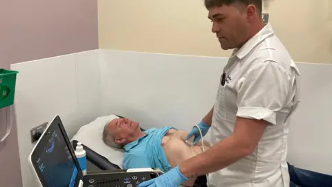 Trevor Johnston lies on a hospital bed while a nurse performs a scan on his abdomen. There is a laptop with an image of his stomach. 