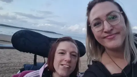 Sarah Lilly Vicky and Sarah pictured at a beach. Vicky is in her wheelchair and Sarah is stood next to her