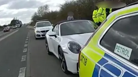 A white Porsche car, pulled over into a layby, with an Essex Police vehicle parked in front and behind it. A man is stood next to the car in a grey jumper with his face blurred out, and two police officers are also stood to the side of the car with their faces blurred out. 