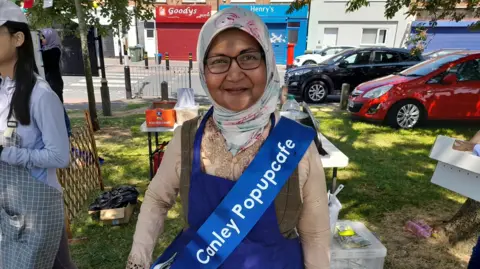 A woman standing in a park on a sunny day, shaded by the threes. She is wearing glasses and a headscarf. Over her clothes she has a blue tabard and a blue sash reading "Canley Popupcafe".