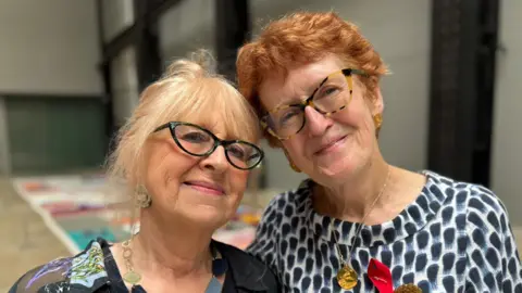 Cathy and her friend Grace smile at the camera. They are both older women wearing glasses and brightly coloured tops and jewellery. They are standing in a hall with parts of the quilt on the floor behind them.
