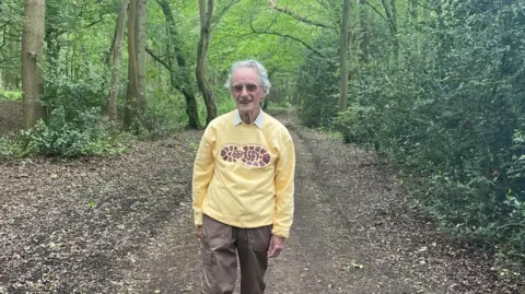 Gerald Drewett An older man with grey hair and dark glasses facing the camera as he walks in woodland. He is wearing a white shirt and a yellow top which has a footprint design on it with the words "a nineties Odyssey" written in the middle.