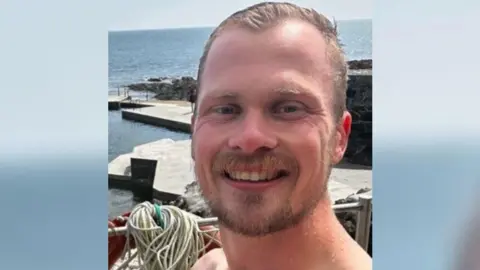 Matt Holland is pictured from the neck up with wet hair. He has a short blond beard with short blond hair. He is smiling at the camera. Behind him is a platform surrounded by water. 