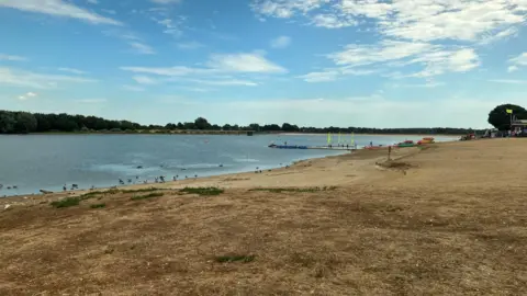 Alton Water is bordered by dried vegetation and dusty ground. There are green trees on the other side of the lake.