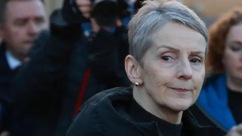 A close-up image of Lady Eleanor Donaldson as she walks out of court. She is an older woman with short, grey hair and has a neutral expression on her face. There are journalists in the background taking videos and pictures of her. Lady Donaldson is wearing a black, puffer raincoat and a pair of silver stud earrings.