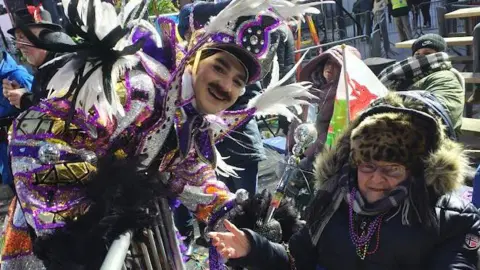 Fiona S-Hedges Photograph of Fiona pictured with one of the Mummers captains, who is dressed in a vibrant purple and gold glittery folk costume. She wears a leopard furry hat, a purple scarf and a black puffer coat. She wears small reading glasses and holds her thumb up to the camera. A captain of the Mummers parade leans down to her and smiles. He wears a vibrant blue, purple and orange sequined costume and has a large red joker smile painted on his face. 