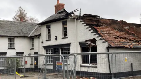 Jill Bennett/BBC A white-painted brick building with a partially collapsed roof. It is surrounded by metal fencing 