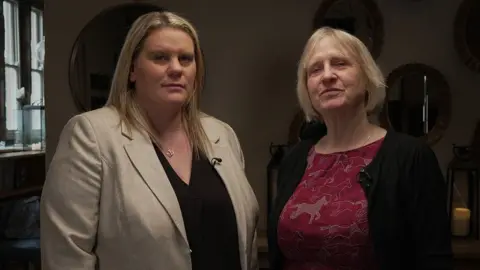 Micala and Sue stand side by side looking defiant. Micala has long blond hair and is wearing a black shirt and light brown jacket. Sue has short blond hair and is wearing a red top with brown horses on it and a black jacket. Behind them is a wall with several mirrors on it