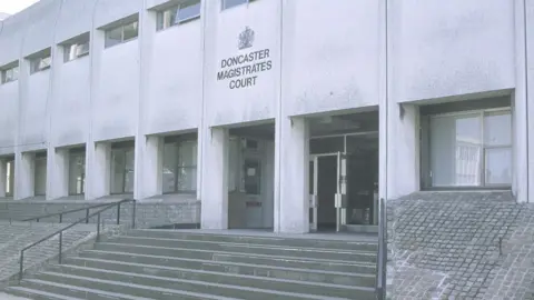 The exterior of a large, concrete building identified by the sign above the entrance as “Doncaster Magistrates’ Court”. The structure has a distinctly utilitarian, modernist design, with tall, rectangular pillars and evenly spaced windows along the upper level. A wide set of steps leads up to the main entrance, which has glass doors, one of which is standing open. To the right of the steps, the ground slopes upward in a textured ramp made of small, square paving stones. Metal railings run alongside the steps.
