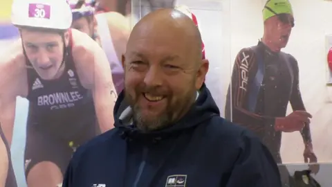 A bald man with a short brown and grey beard wearing a blue waterproof jacket stands, smiling, in front of a wall showing images of the Brownlee brothers competing in triathlons. 