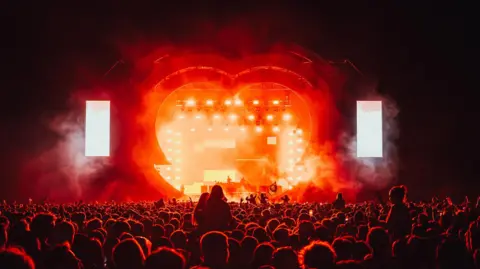 A heart-shaped stage at night with orange lights and stage smoke, with the silhouettes of a huge crowd in front.