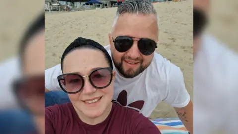 Ed Growden Ed and his wife Kerry sitting on a beach smiling. Kerry is sat on the left and is wearing a maroon tshire, sunglasses, and has dark tied-back hair. Ed is on the right, has grey hair, is wearing sunglasses and a white tshirt.