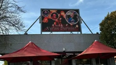 A large advertising screen situated above a coffee shop in Dundalk town centre. It shows several photos of the missing boy with the words: "Where's wee Kyran Durnin?"