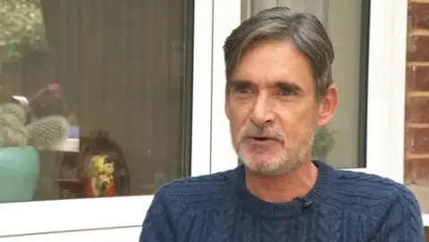 Gary Webster sits outside in front of a white framed window. He has short light brown hair with a little grey, he also has a white stubbly beard. He is wearing a navy blue cable knit crew neck jumper