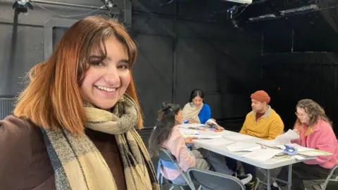 A woman with brown hair and a yellow checked scarf with four people sitting around a table n the background