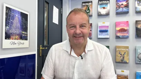 Andrew Earle sits in front of his computer in his travel agents in Hull city centre wearing a white shirt with holiday brochures to various destinations in the background.