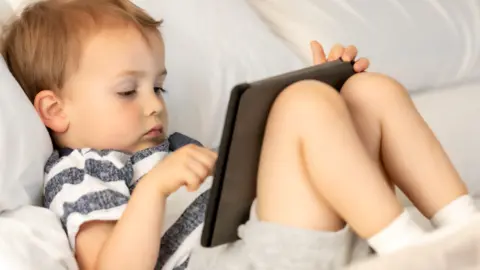 Getty Images A toddler lying on his back in front of a screen, wearing grey shorts and a blue and white stripey tshirt. 